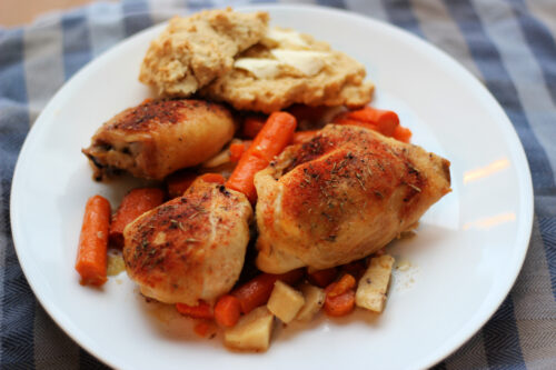 Plated chicken thighs on top of carrots and parsnips with biscuit in background