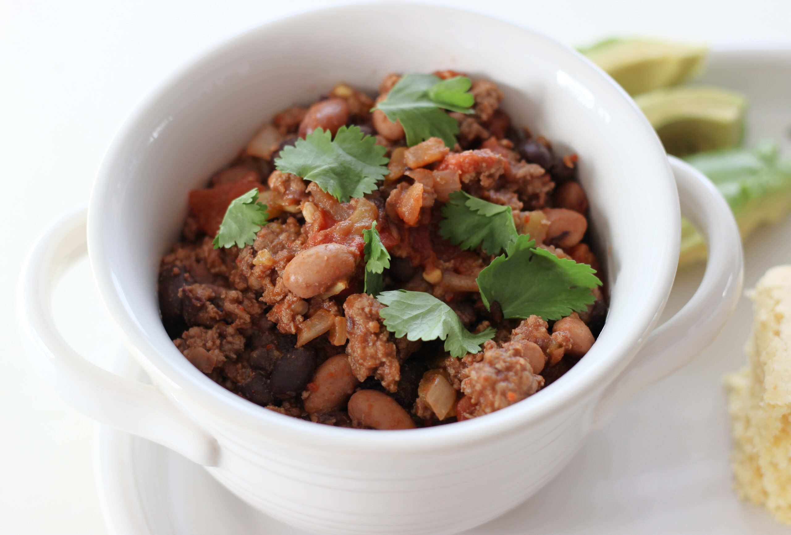 Chili in a bowl with cilantro next to corn bread and avocado