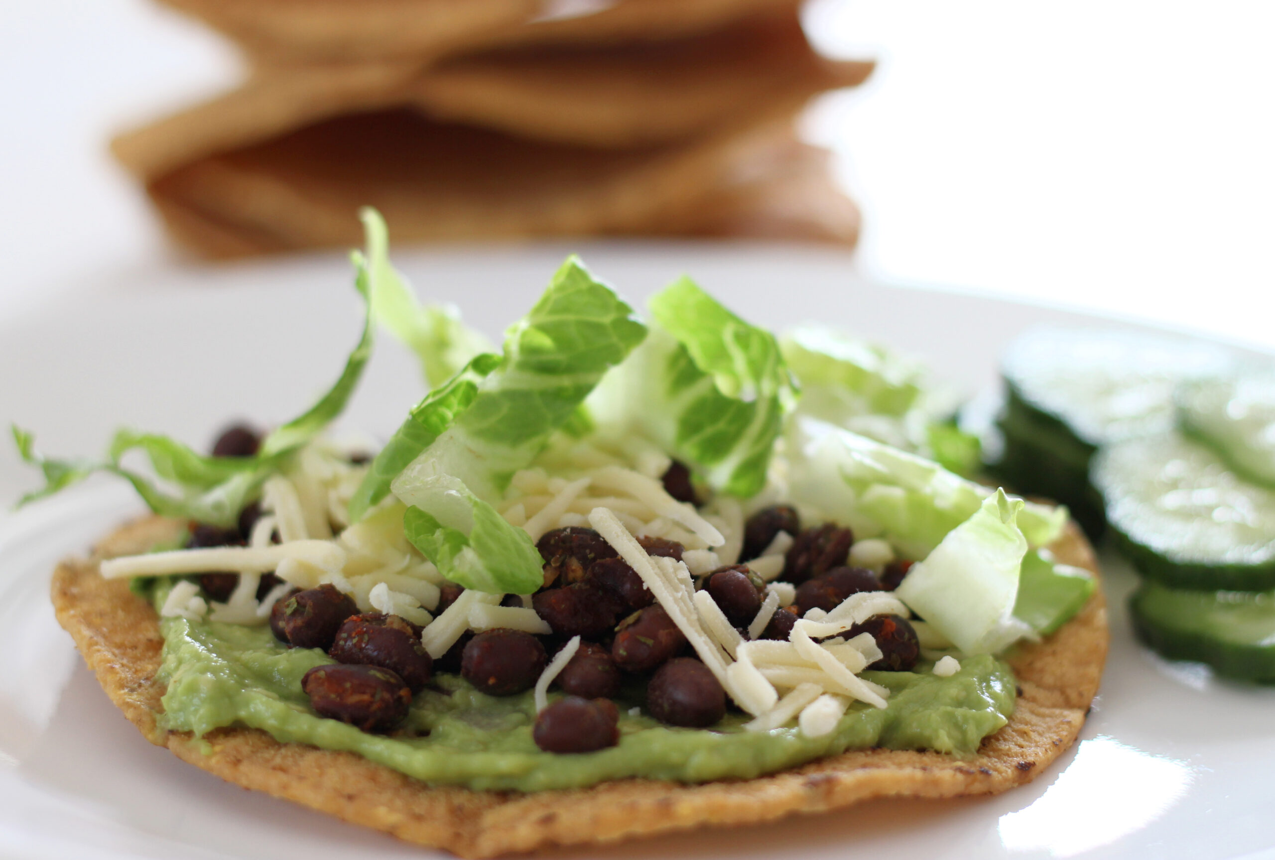 Black bean tostada with guacamole on a plate with cucumbers and tostadas in the background
