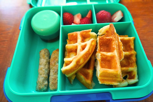 Waffles packed with syrup, chicken sausage links and sliced strawberries